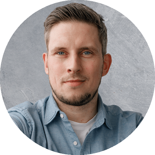Felix, a professional speaker with short light brown hair and a trimmed beard, wearing a light blue denim shirt over a white t-shirt, in front of a textured gray background.
