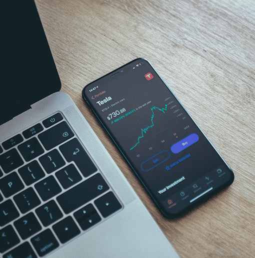 A smartphone displaying a stock trading app with Tesla stock price and performance chart, placed next to a laptop on a wooden desk.